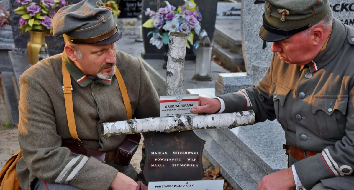 Zdjęcie przedstawia moment przykręcania pamiątkowej tabliczki do krzyża na grobie powstańca wielkopolskiego